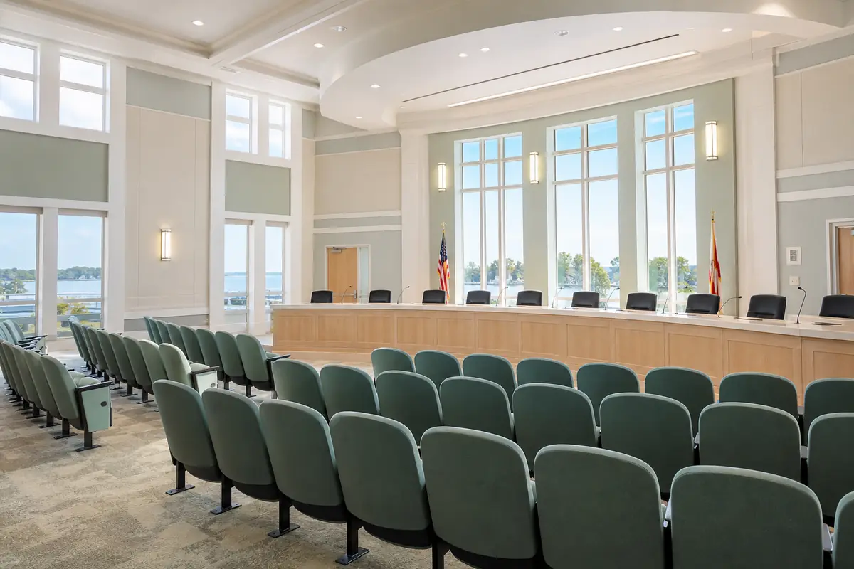 An illustration: rows of empty public seats facing a long curved dais in a bright county meeting room with tall windows