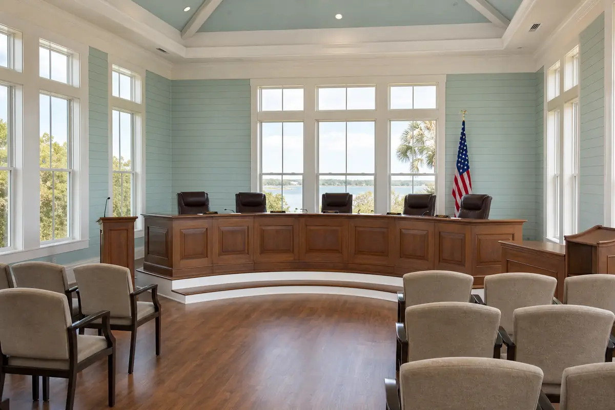 An illustration: a small-town city council chamber with a wooden dais, empty leather chairs and a flag, soft daylight through tall windows