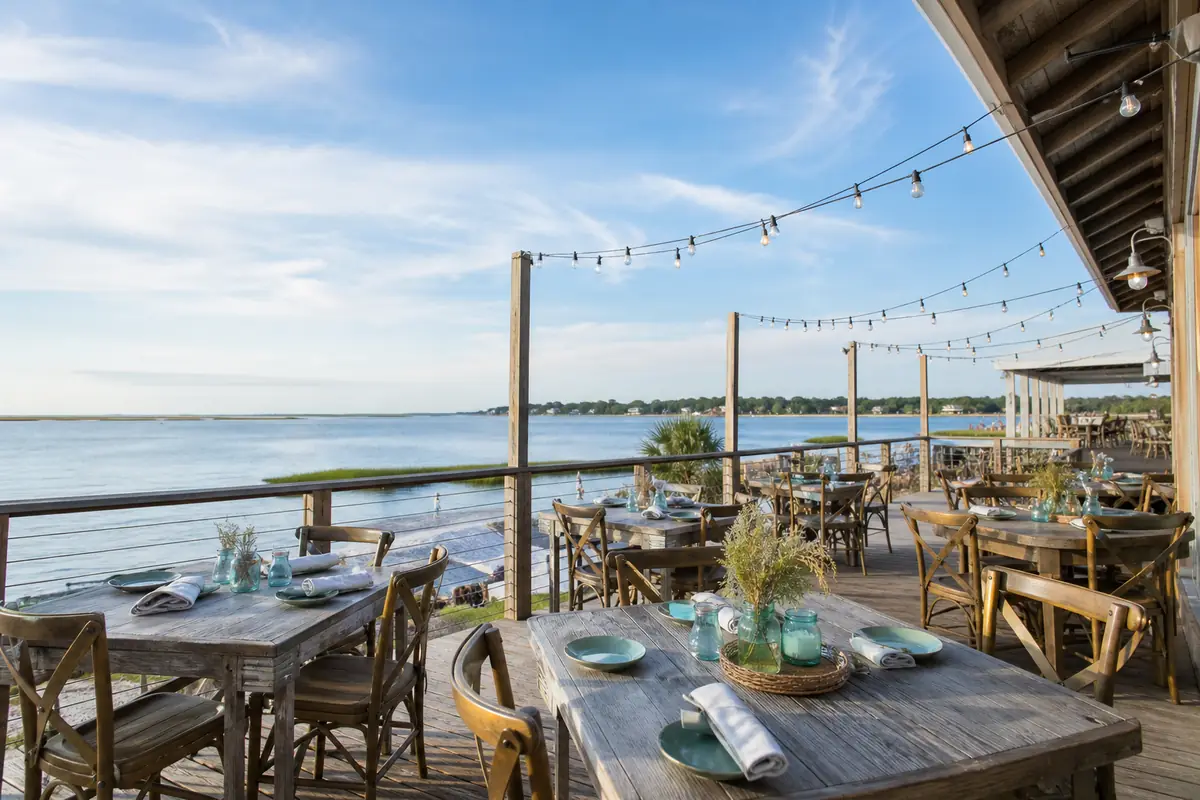 A relaxed coastal restaurant's outdoor deck with wooden tables set for a meal, string lights, and the water beyond: an illustration for Freeport's eat tile.
