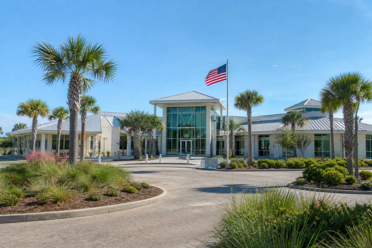 An illustration: a modern county government building with a glass entrance, a flag and landscaped palms under a blue sky
