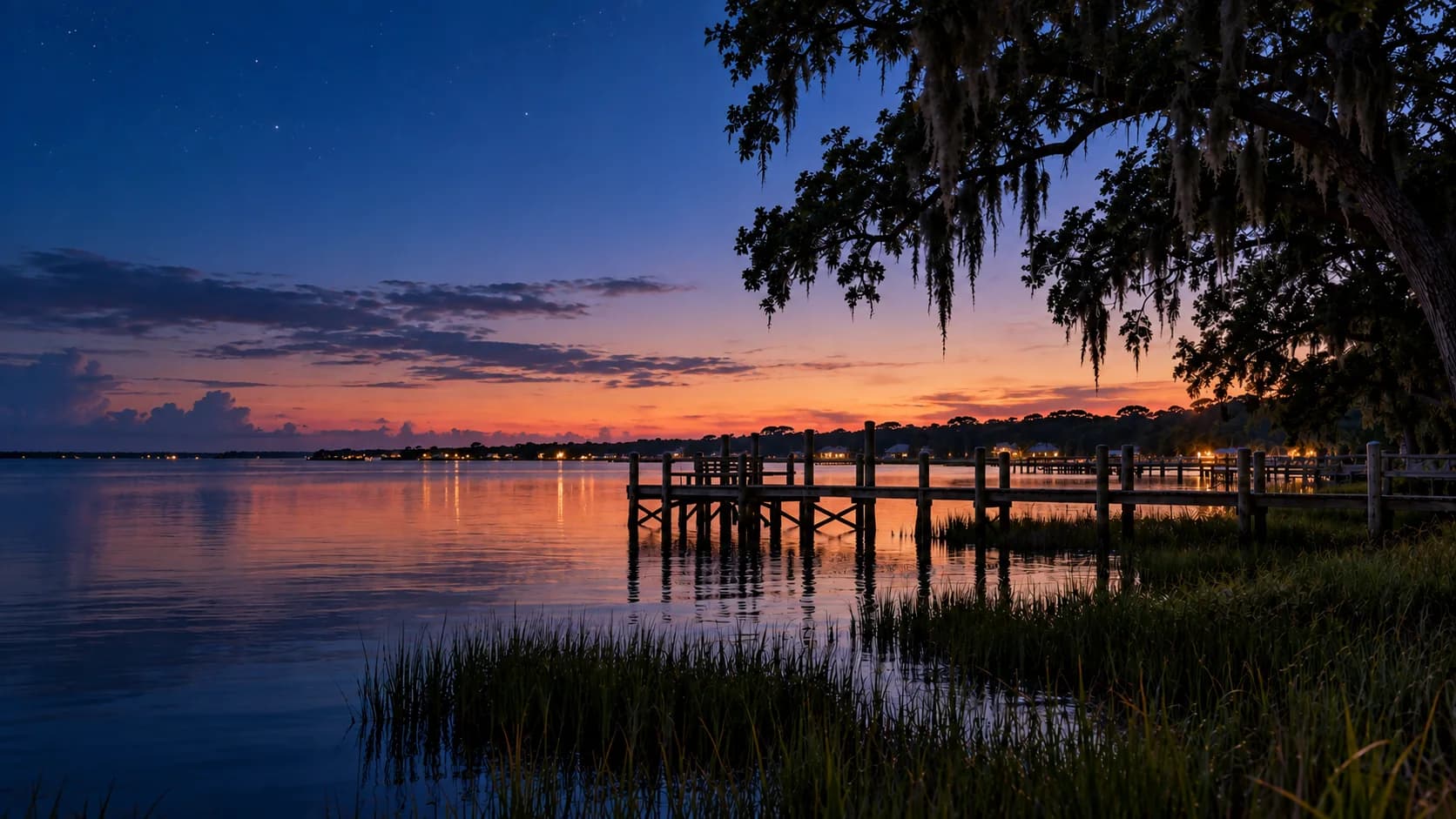 Choctawhatchee Bay at Freeport after sunset: the last orange light on the horizon, shore lights reflected on still water, and a dock under a mossy live oak.